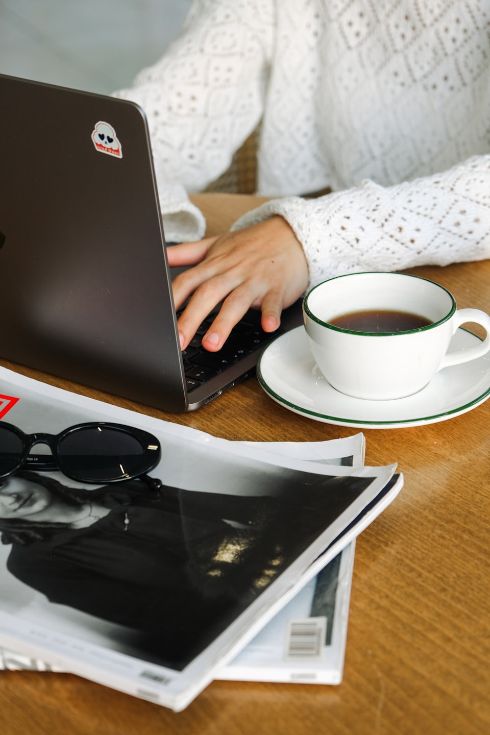 Hands typing on a laptop next to a coffee cup and magazines.